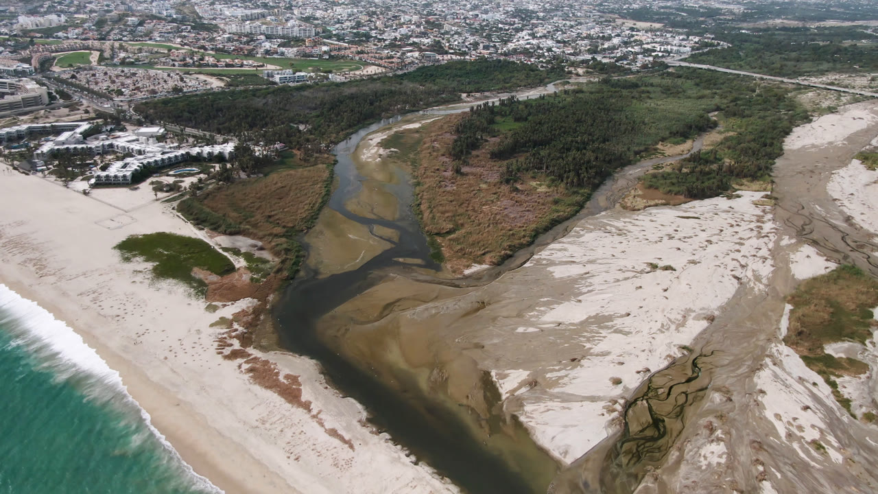 High aerial frame in San Jose del Cabo, Mexico, showing the river bend centered between pale sand banks, green vegetation, white resort buildings, and a strip of ocean surf, with no visible people.