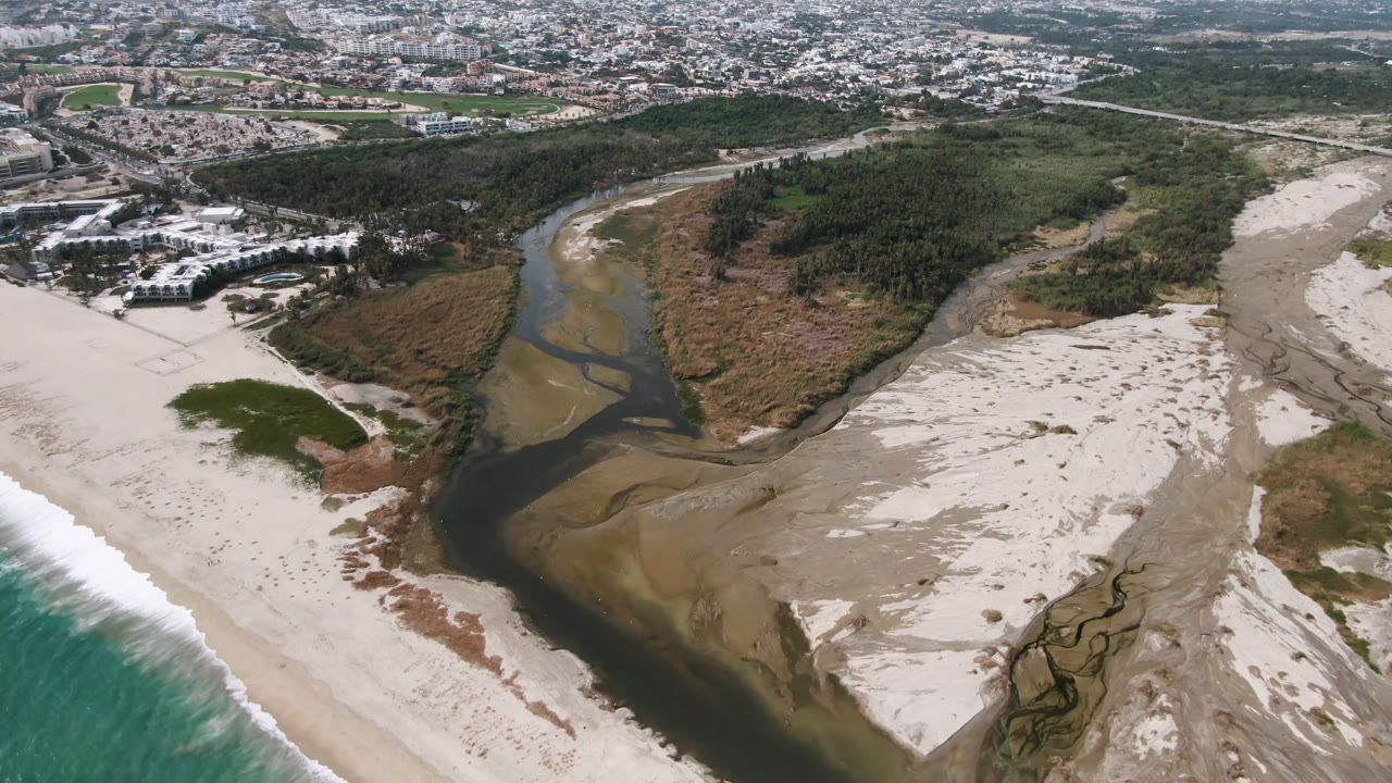 Aerial coastline view with a river cutting through sand toward the ocean.