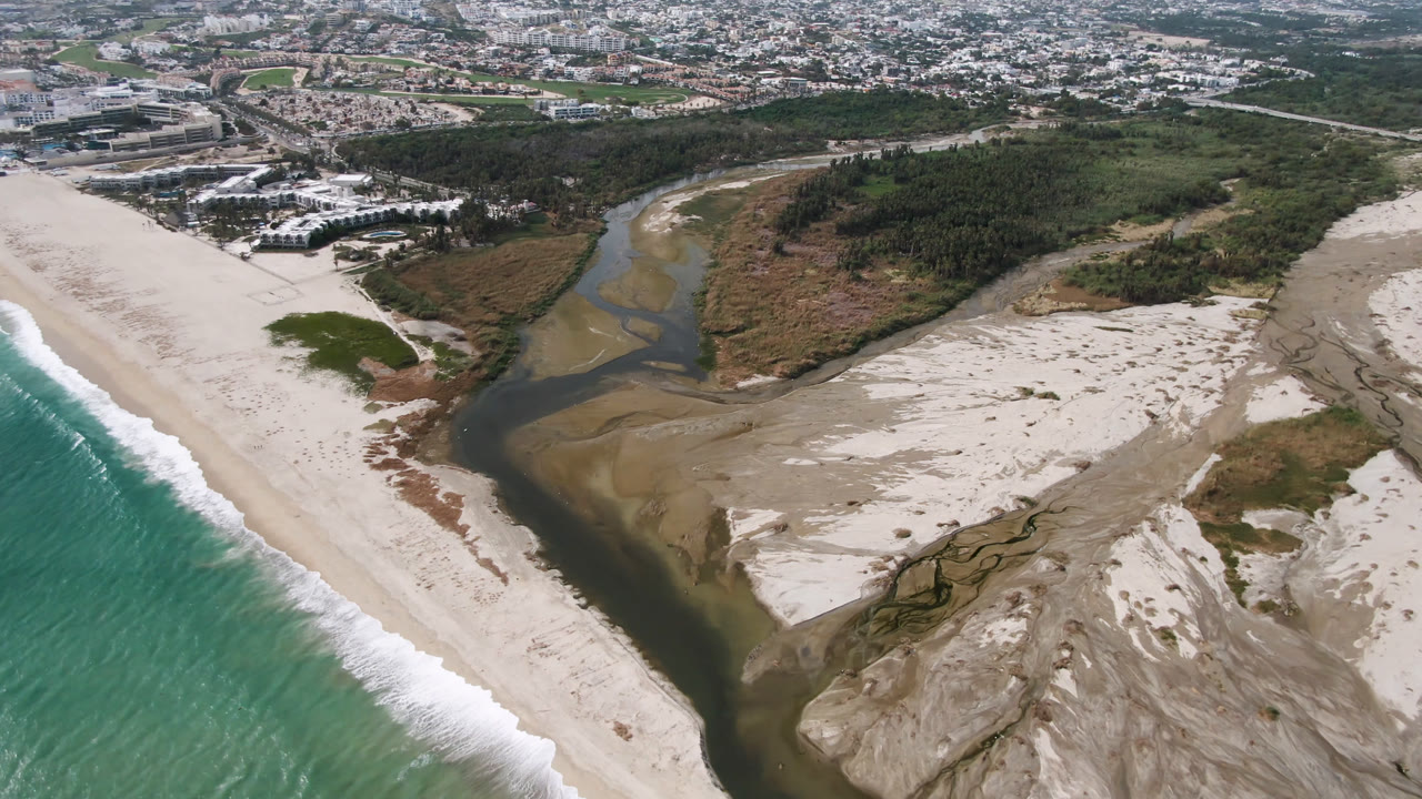 Aerial view over San Jose del Cabo showing turquoise surf at left, a pale beach, a dark river channel through sand banks, inland vegetation, and no visible people.