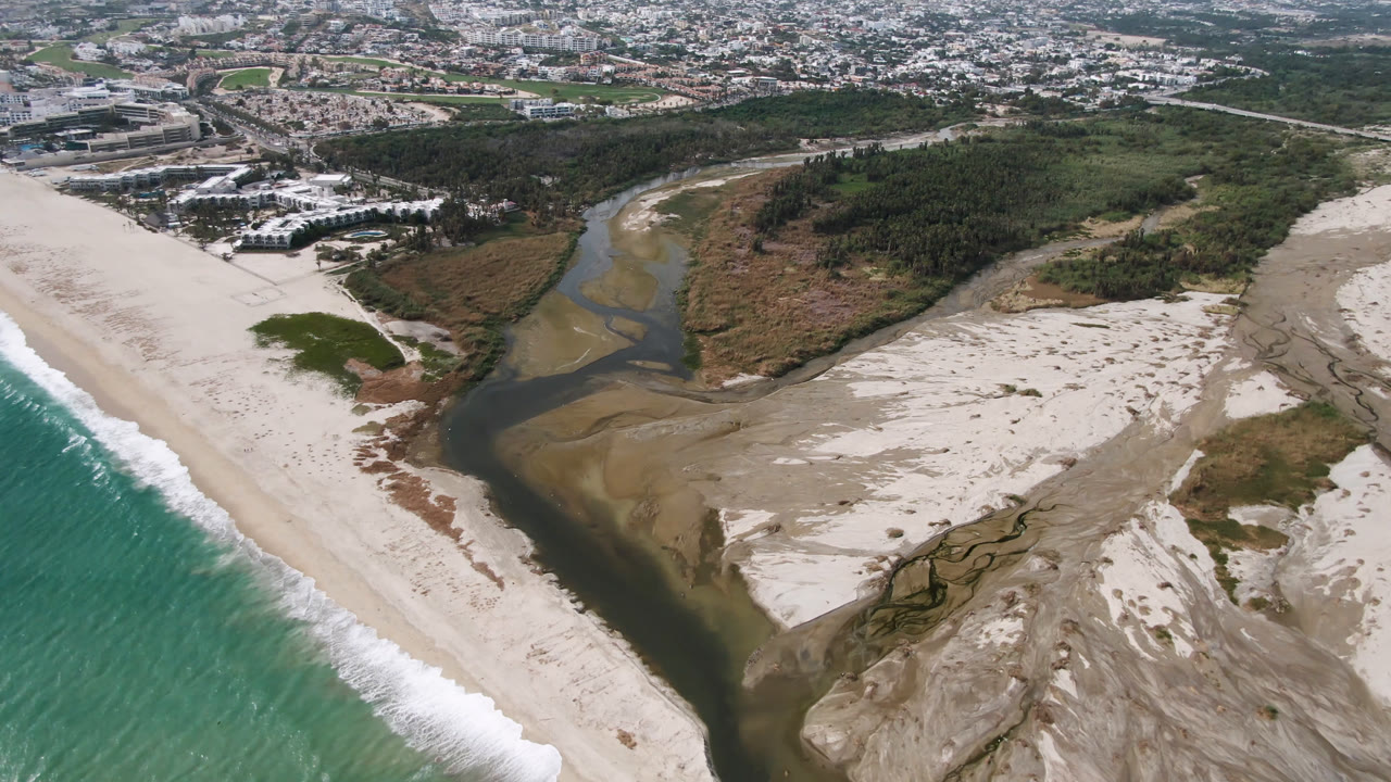 High aerial frame in San Jose del Cabo with surf running along the right side, a winding river through light sand bars, green brush, buildings, and no visible people.