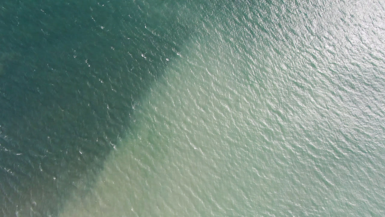 Overhead view of blue green ocean in San Jose del Cabo, Mexico, with darker teal water across the upper frame and pale shallow color below.