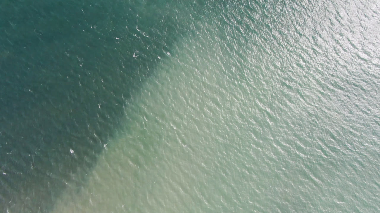 Aerial top-down water view in San Jose del Cabo, Mexico, where pale green shallows meet a darker teal band under bright surface glare.