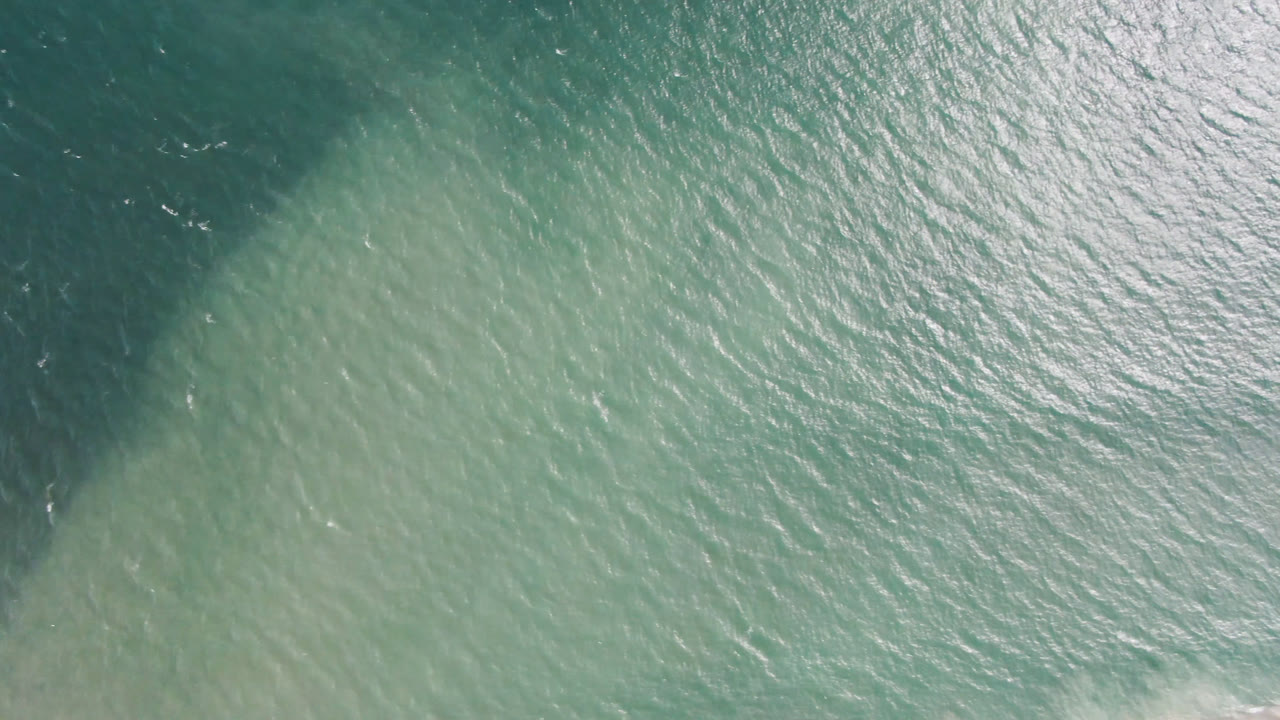 Aerial frame over San Jose del Cabo water with green-blue ripples, a darker area at the left edge, and no visible people.