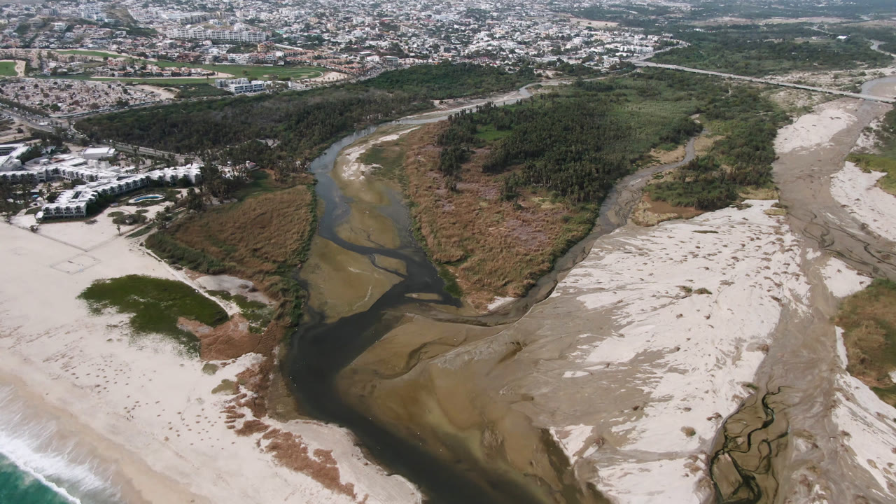 Aerial view of a dark river channel crossing pale sand toward the surf.