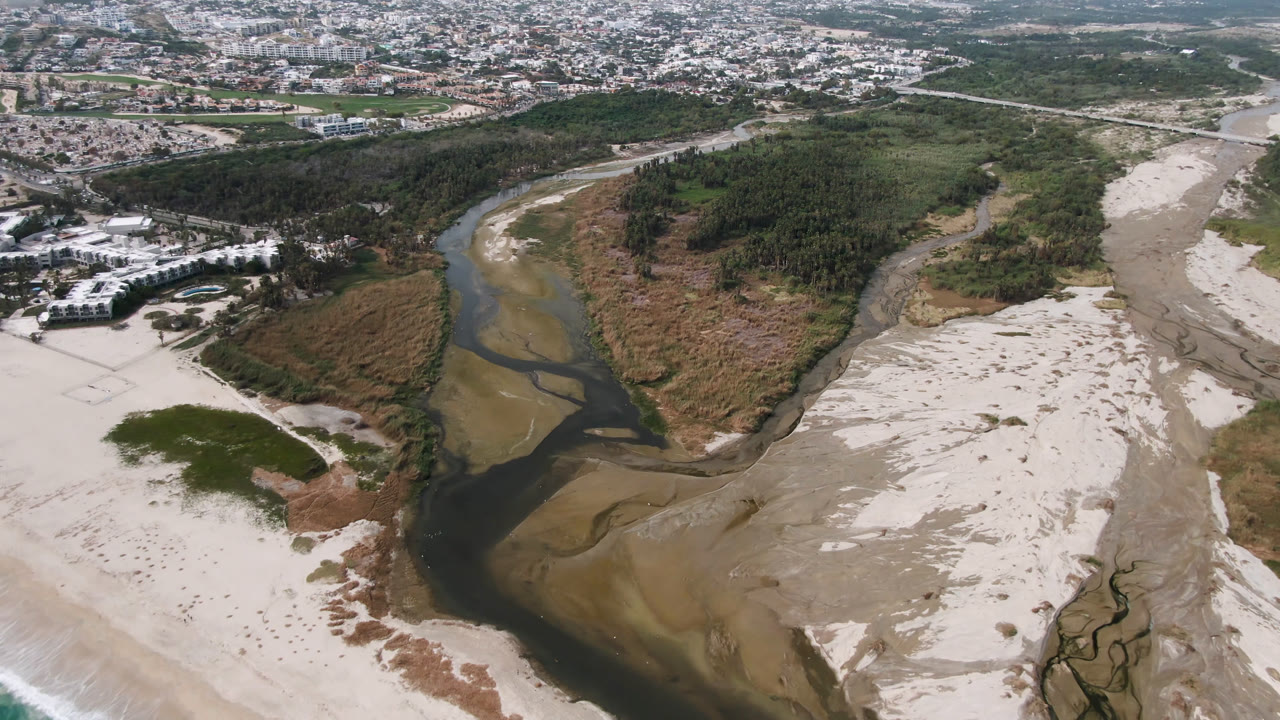 Aerial frame from San Jose del Cabo with the river channel centered beside tan sandbanks, a broad pale beach to the right, resort buildings near the left edge, distant city blocks, and no visible people.