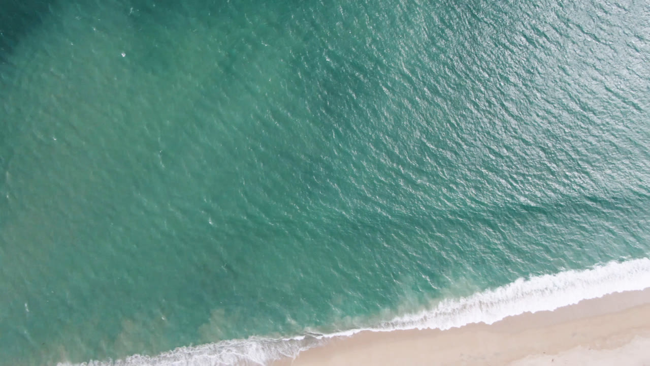 Overhead view of teal water and a narrow pale sand beach in San Jose del Cabo, with white surf just reaching the shoreline and no visible people.