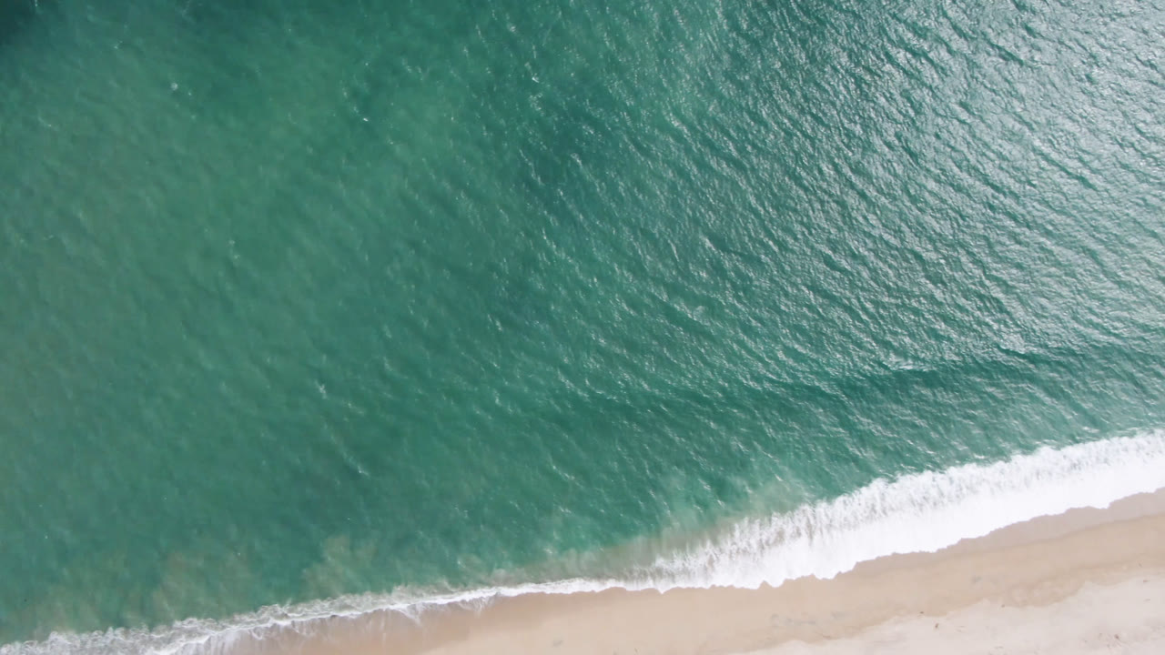 Aerial view over San Jose del Cabo shoreline where a white wave line spreads diagonally across pale sand, with open teal water above and no visible people.