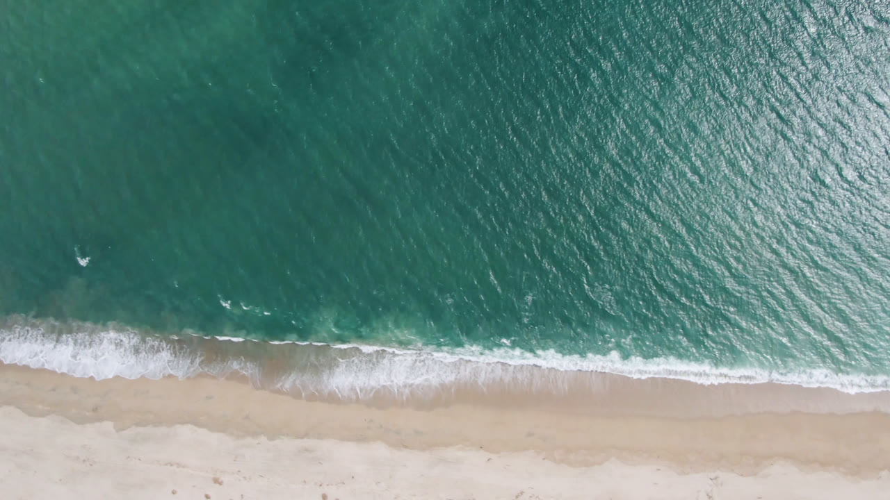 Overhead view of turquoise ocean meeting pale sand in San Jose del Cabo, Mexico, with a narrow white surf line and no visible people.