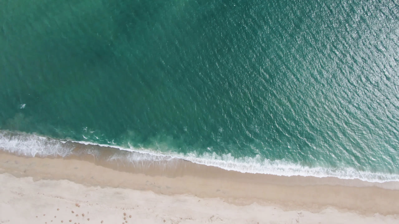 Aerial frame over San Jose del Cabo shoreline with rippled green-blue water, a thin breaking wave, pale beach, and no visible people.