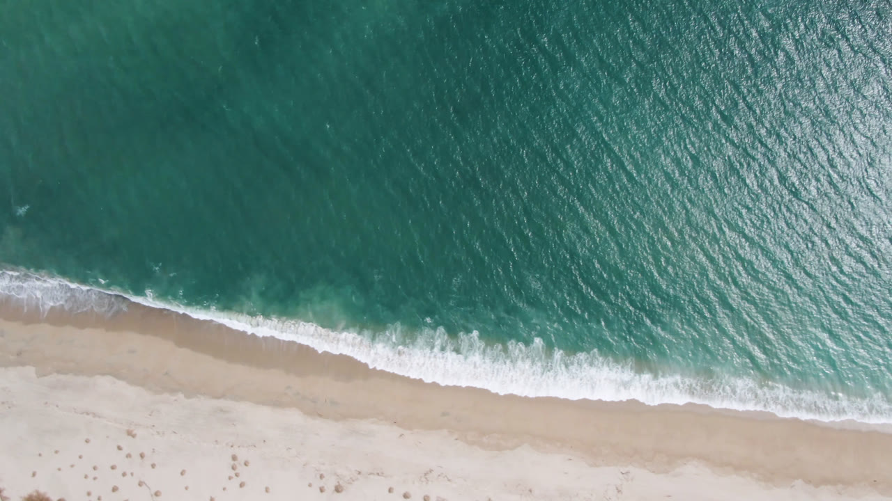High overhead view in San Jose del Cabo, Mexico, with a curved white surf band crossing between teal water and smooth sand, with no visible people.