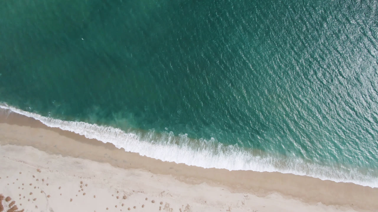 Top-down San Jose del Cabo beach frame with darker teal water above, foam spreading across the shoreline, pale sand below, and no visible people.