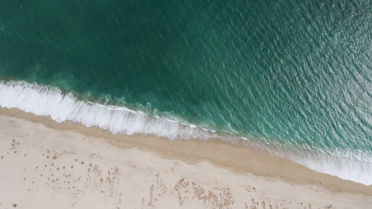 Aerial frame over San Jose del Cabo shows the breaking wave line shifted along the tan shoreline, with textured teal water above and no visible people.