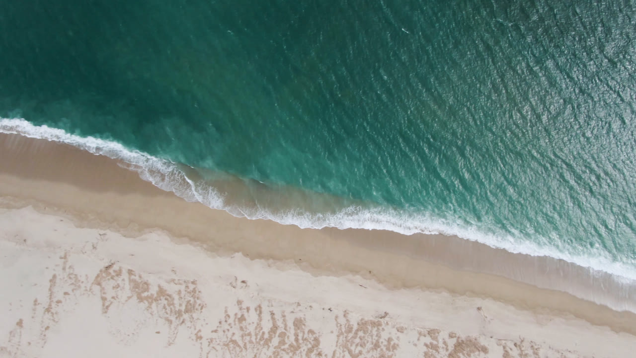 A diagonal band of white foam crosses the San Jose del Cabo shoreline as shallow water washes onto empty sand, with no visible people.