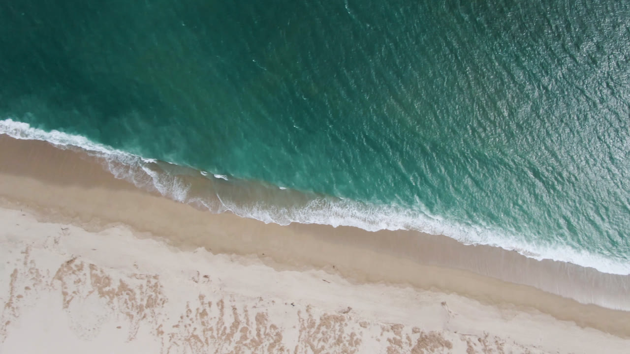 Turquoise water breaking along a pale beach from above.