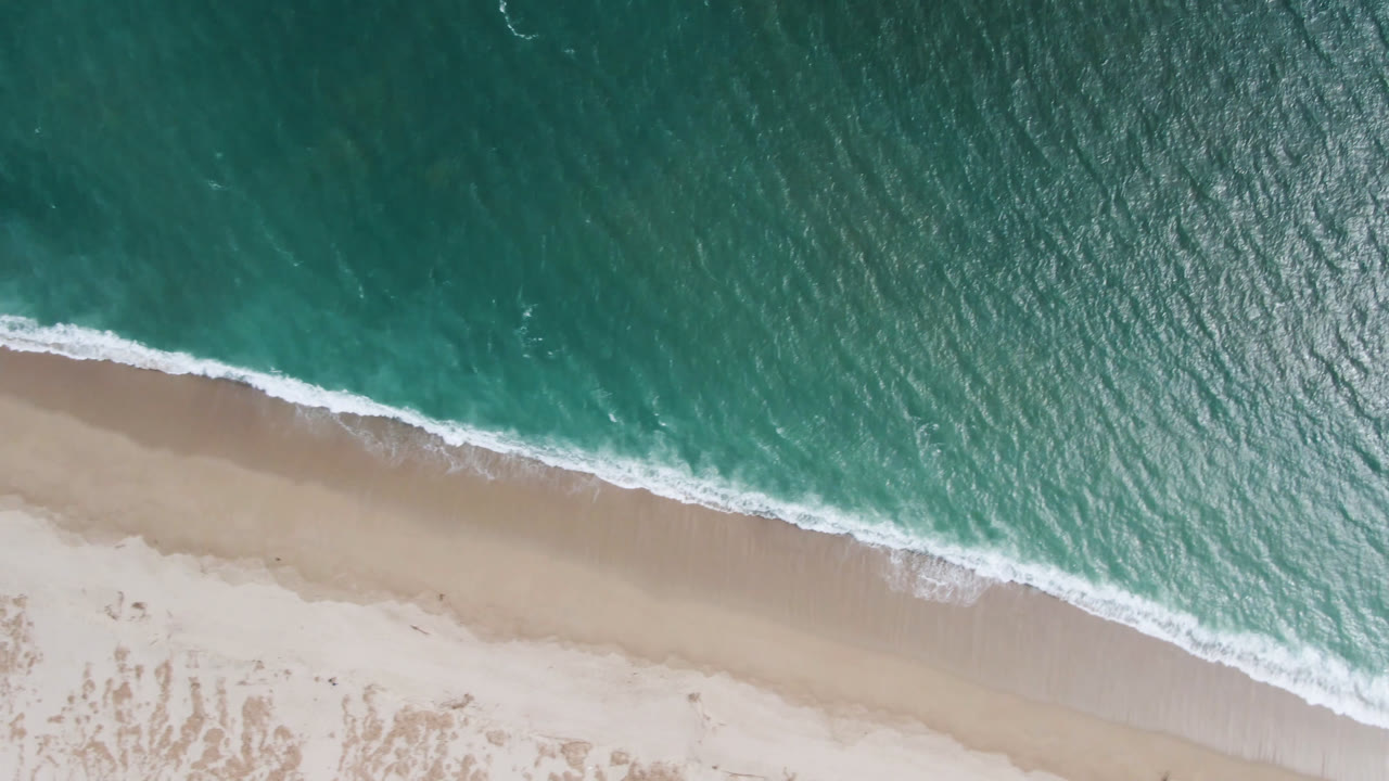 Top-down aerial frame from San Jose del Cabo showing turquoise water, a diagonal line of white surf, pale sand, and no visible people.