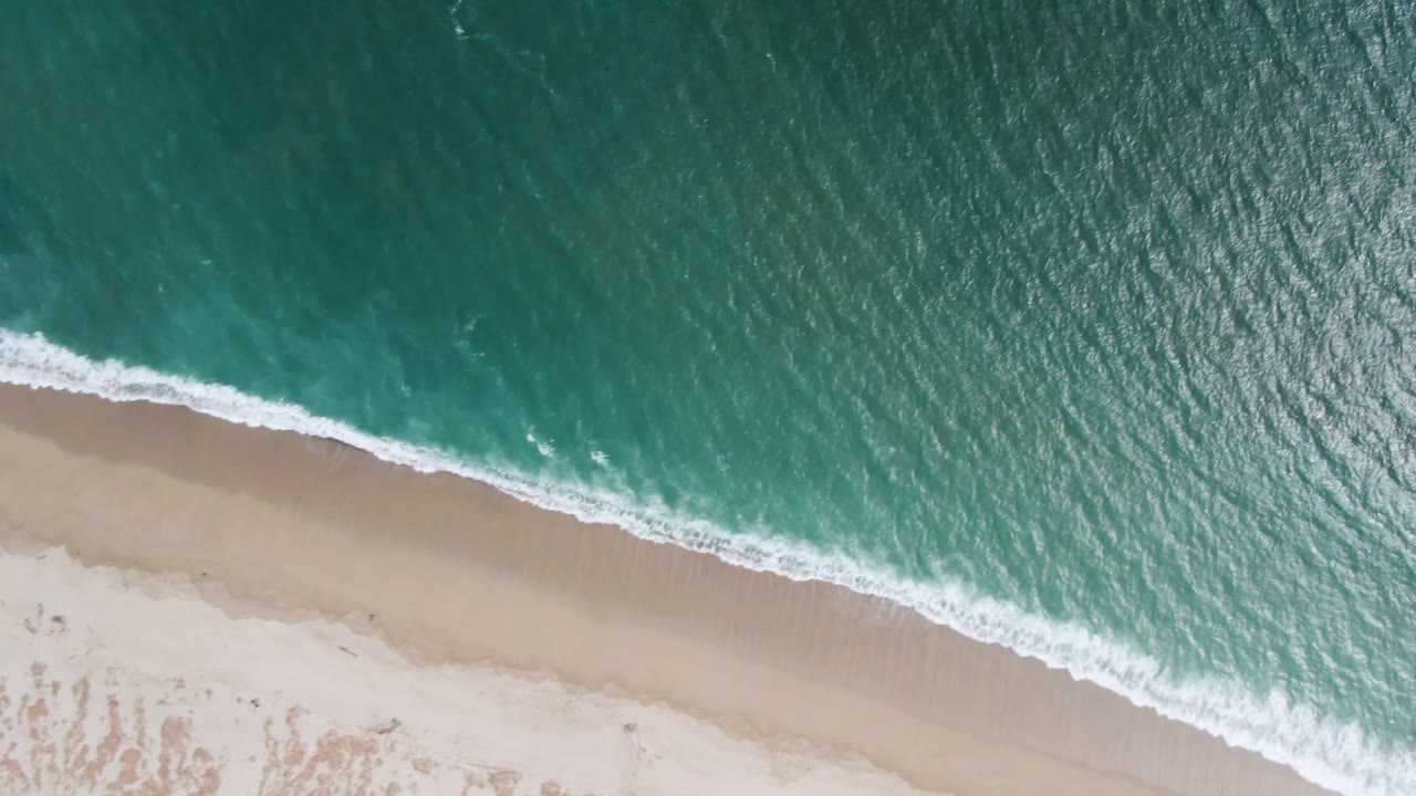 San Jose del Cabo aerial frame with the foamy wave edge slightly farther along the diagonal shore, turquoise water texture, pale sand, and no visible people.