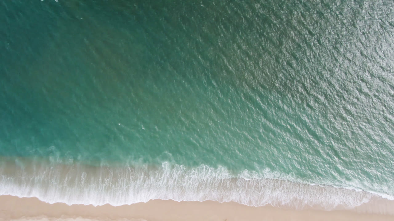 Top-down aerial view of turquoise water and white surf reaching pale sand in San Jose del Cabo, Baja California Sur, with no visible people.
