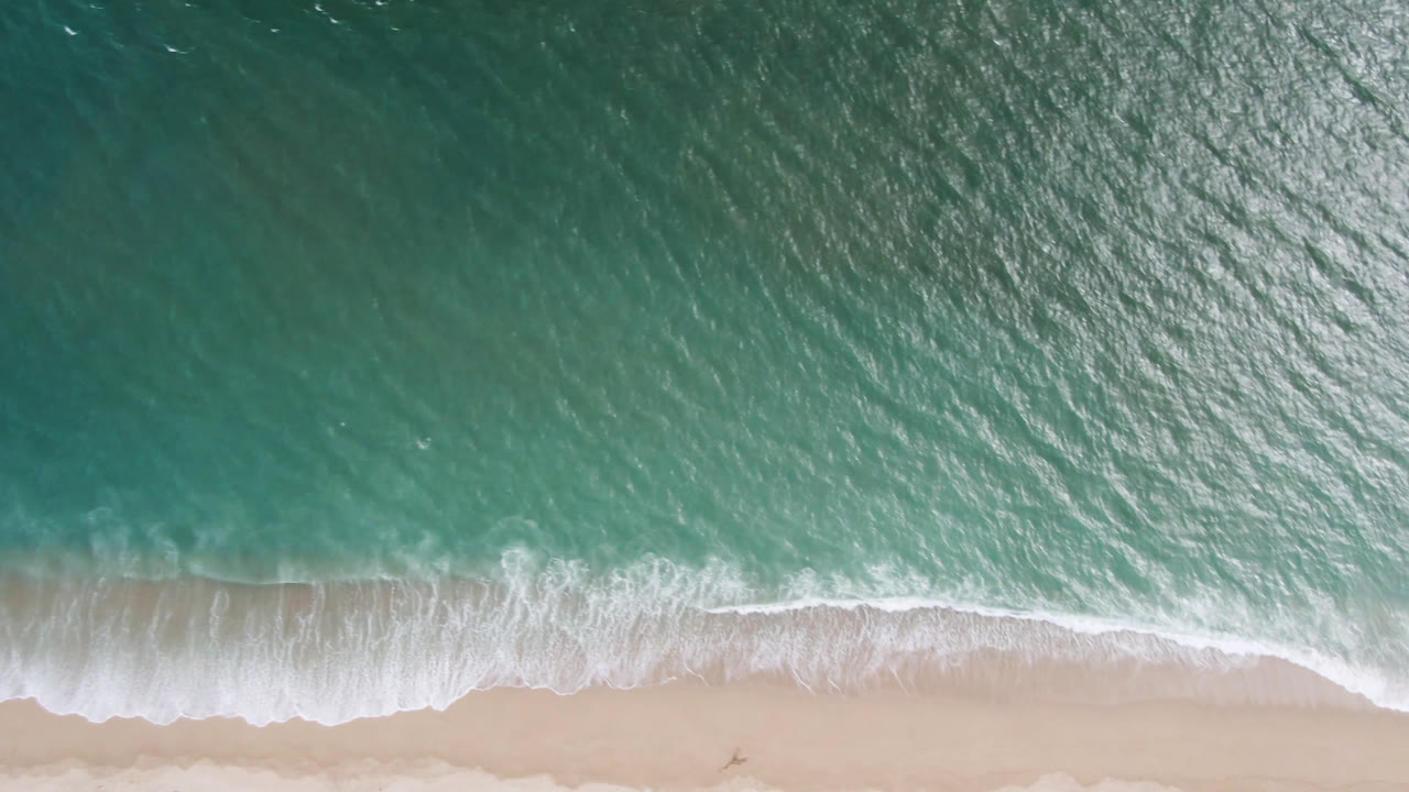 Aerial frame over San Jose del Cabo surf with white foam spread wider across the sand and clear green-blue water above it, with no visible people.