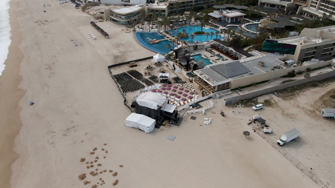Overhead frame of a San Jose Del Cabo beach resort with pools, palm-lined landscaping, stage structures, lounge seating, vehicles on sand, and no visible people.