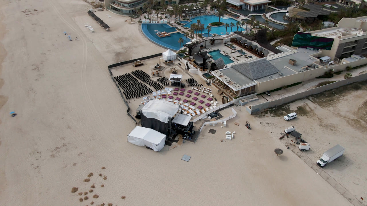 High aerial view in San Jose Del Cabo showing a resort pool area, rows of dark chairs, purple seating blocks, white loungers, tents, service vehicles, and no visible people.