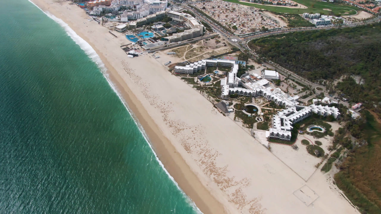 Aerial frame over San Jose del Cabo showing turquoise water at left, a diagonal sandy beach, white resort buildings with pools inland, and no visible people.