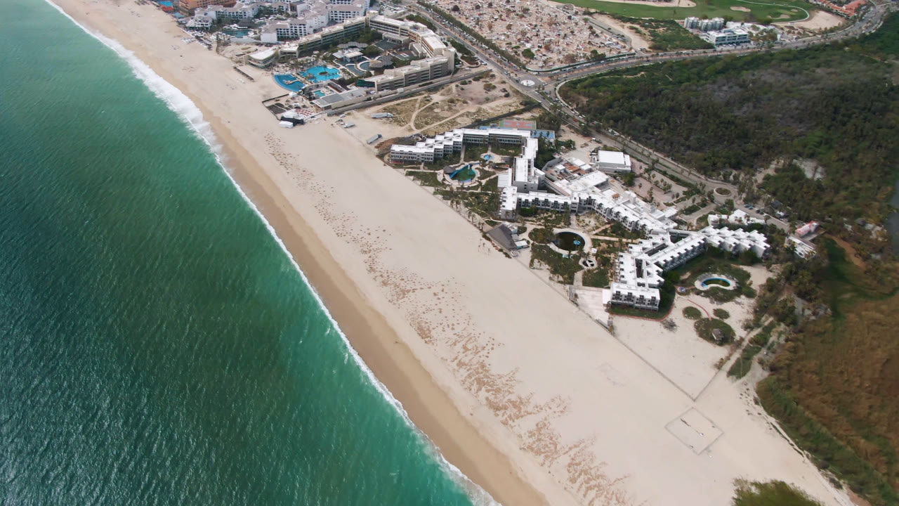 Aerial frame over San Jose del Cabo with the beach stretching diagonally, resort pools near the center, ocean along the left edge, inland roadways above, and no visible people.