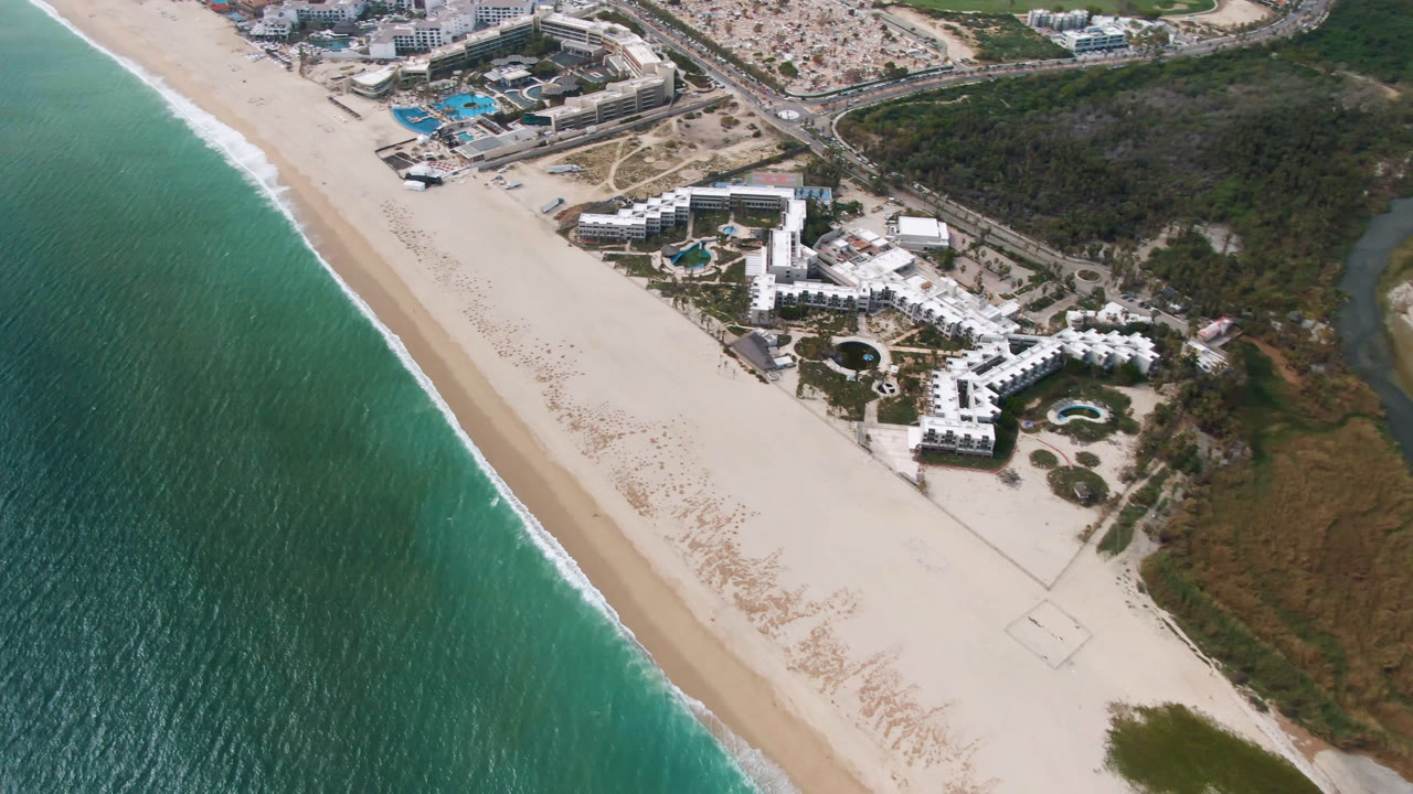 High aerial of white resort buildings beside a broad beach and green-blue water.