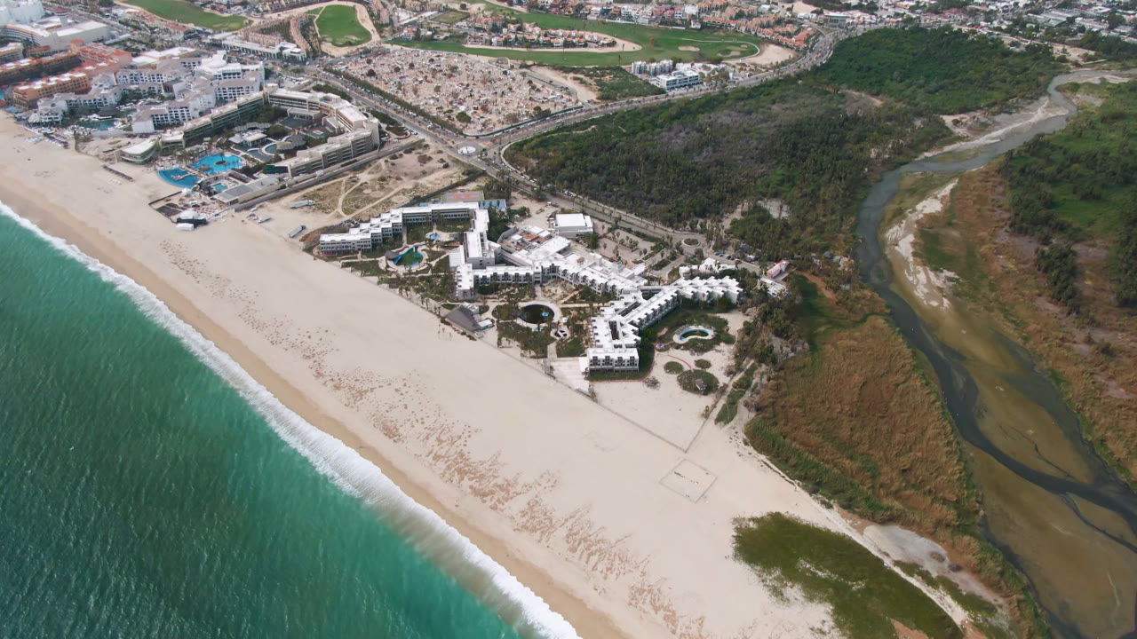 Second aerial frame in San Jose del Cabo with the diagonal shoreline at left, resort pools near the beach, brown-green lagoon water to the right, and no visible people.