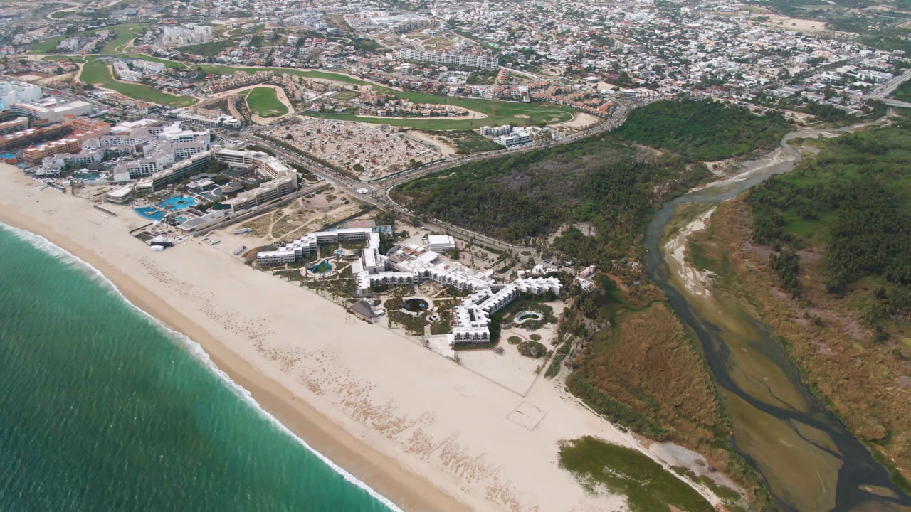 Aerial frame over San Jose del Cabo with the surf line slightly farther along the beach, resort pools near center, the river channel at right, city blocks beyond, and no visible people.