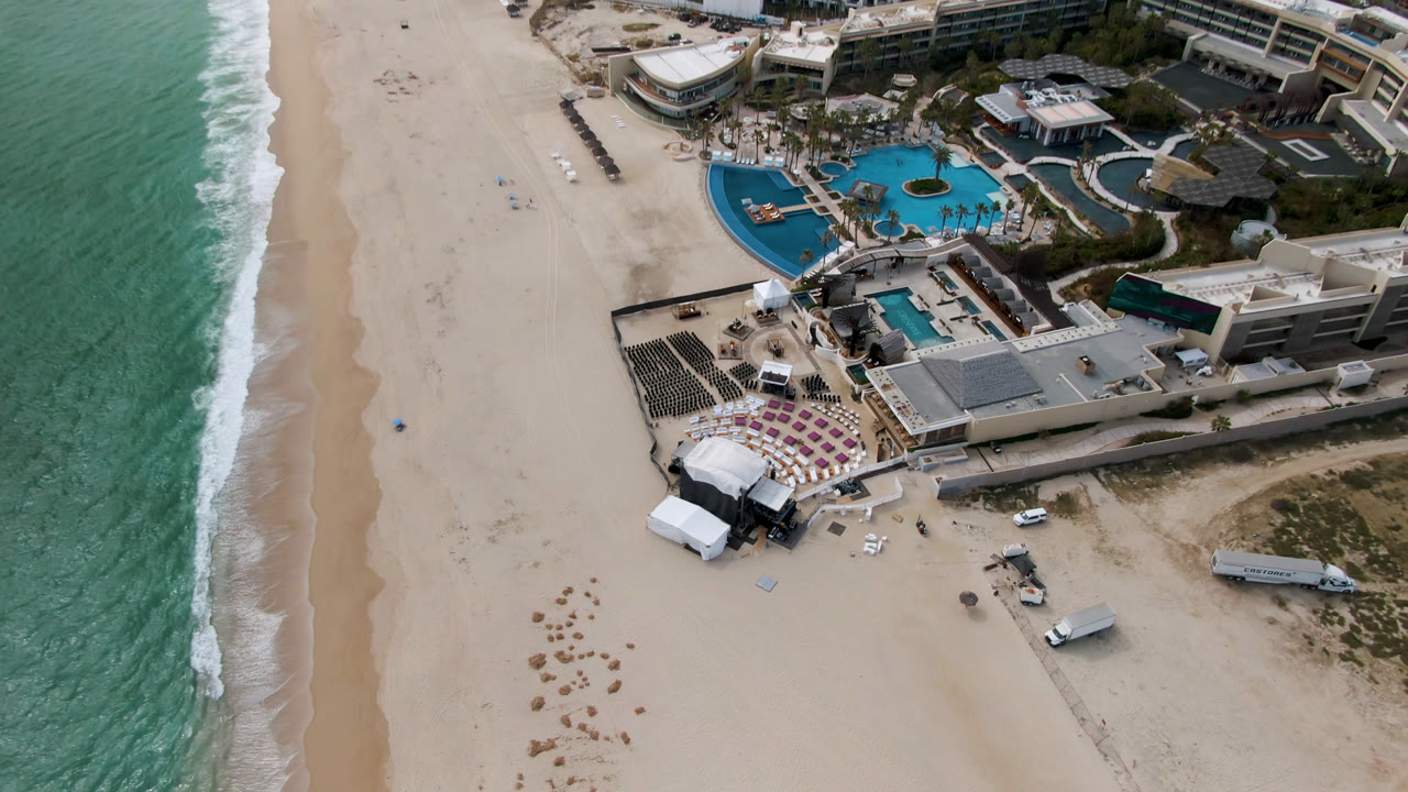 Aerial frame over San Jose Del Cabo showing turquoise surf, a sandy beach, resort pools, white tents, seating rows, and no visible people at the start of the clip.