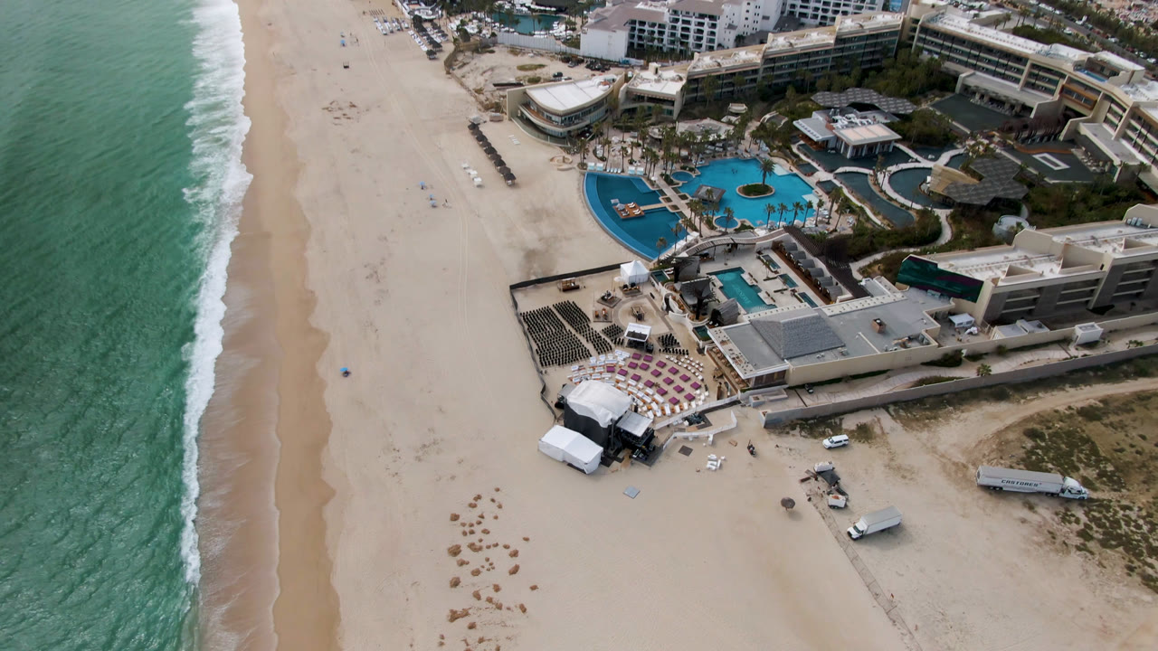 Aerial view in San Jose Del Cabo with the beach running vertically through frame, ocean surf on the right, resort pools and event seating near the lower center, and no visible people.