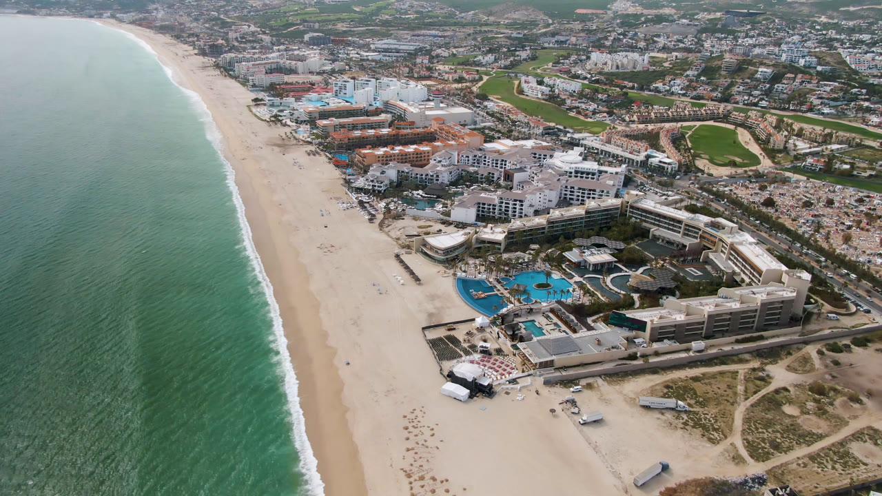 Aerial frame over San Jose del Cabo showing green ocean water, a long sand beach, a beachfront resort with blue pools, roads, and a golf course beyond, with no visible people.