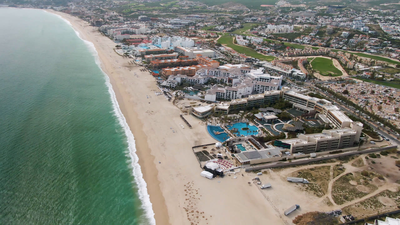 Second aerial frame in San Jose del Cabo with the surf line slightly farther along the left edge, the resort pools centered lower in view, and the golf course and city blocks behind, with no visible people.