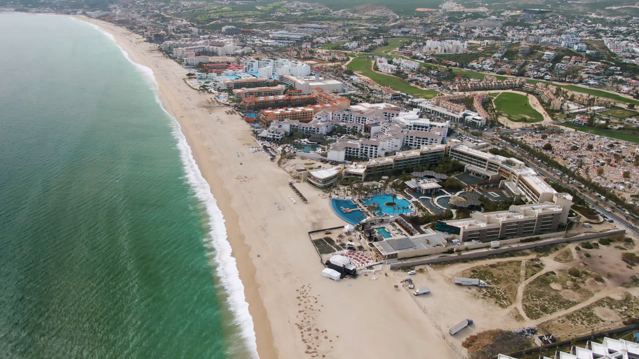 High coastal view of ocean, beach, resort pools, hotel roofs, roads, and nearby golf greens.