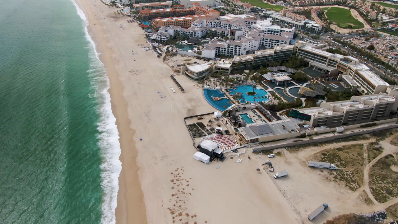 Opening aerial frame over San Jose del Cabo, Mexico, with green-blue ocean at left, a wide sand beach, resort buildings and bright pools at right, and no visible people.