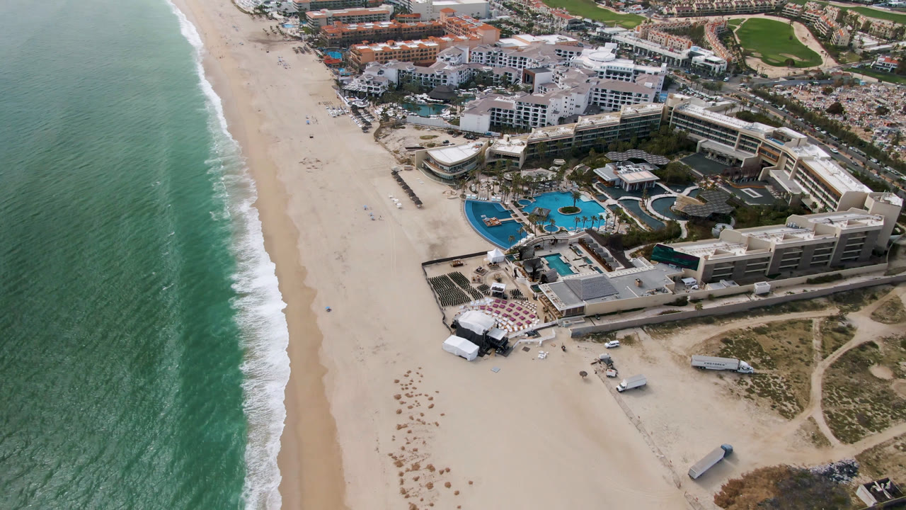 Aerial frame in San Jose del Cabo showing the surf line closer to the left edge, the empty-looking beach through the center, resort pools on the right, and no visible people.
