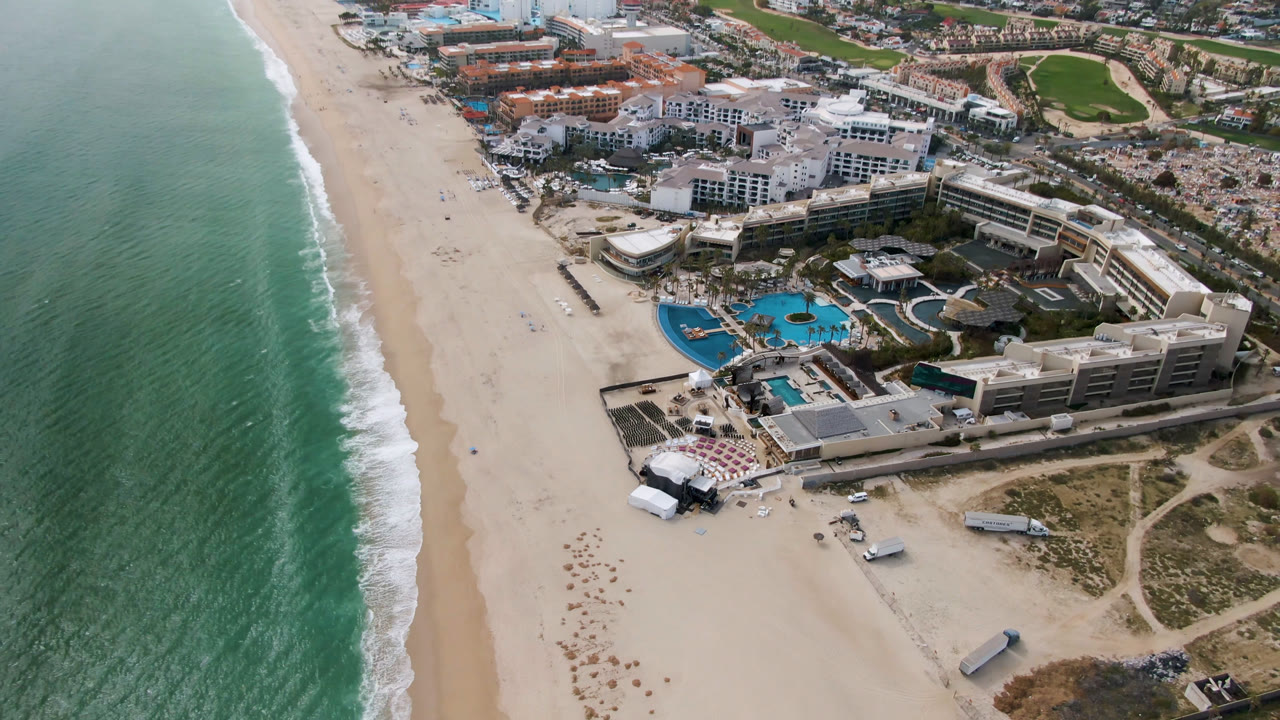 Aerial coastal resort view with ocean, sandy beach, pools, and golf course inland.