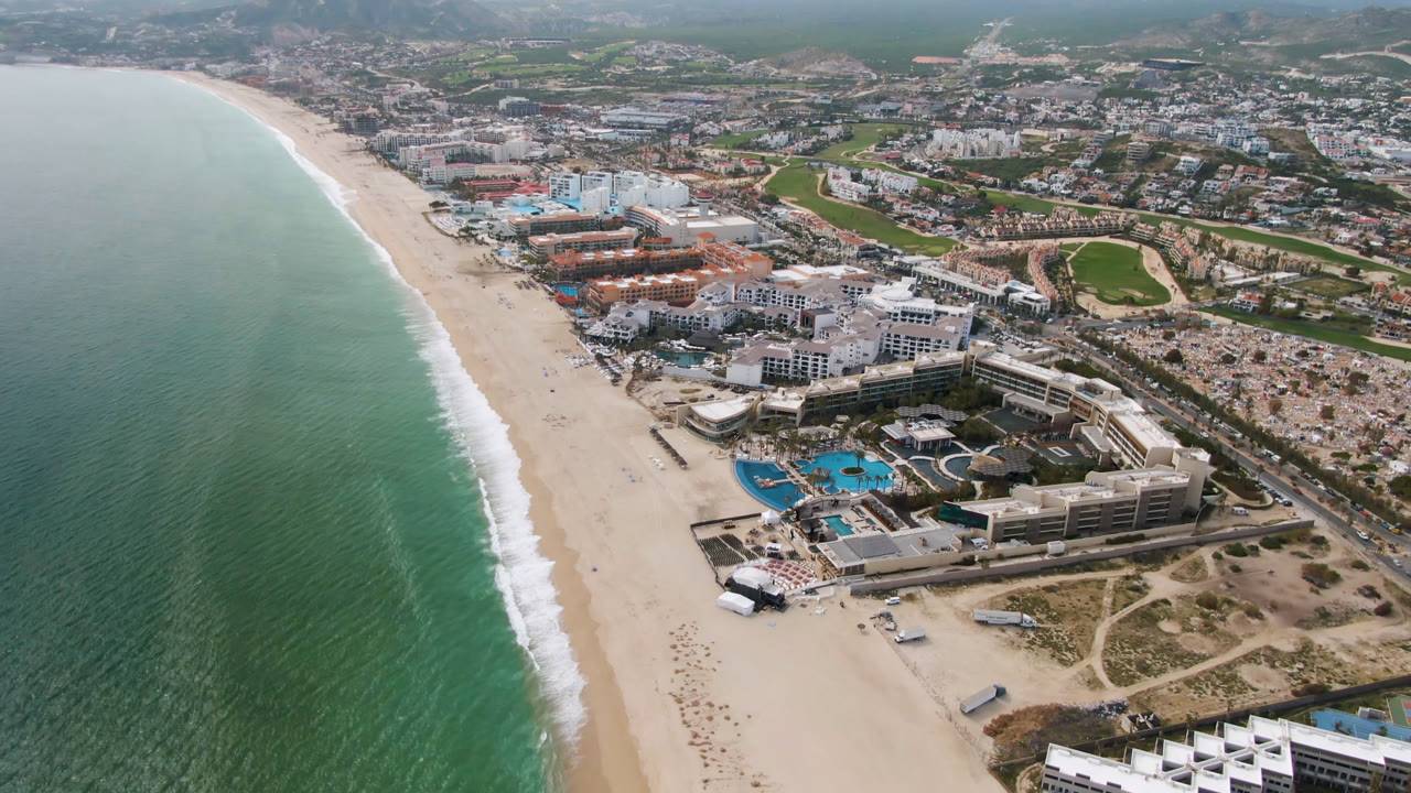 Opening aerial frame over San Jose del Cabo with green-blue ocean on the left, a wide beach, resort pools near the shore, inland town blocks, golf course greens, hazy mountains, and no visible people.