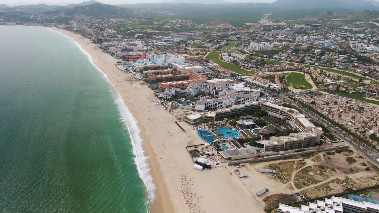 Second frame of the San Jose del Cabo aerial holds the resort strip beside the beach, with blue pools and low-rise buildings in the foreground, the town and golf course beyond, and no visible people.