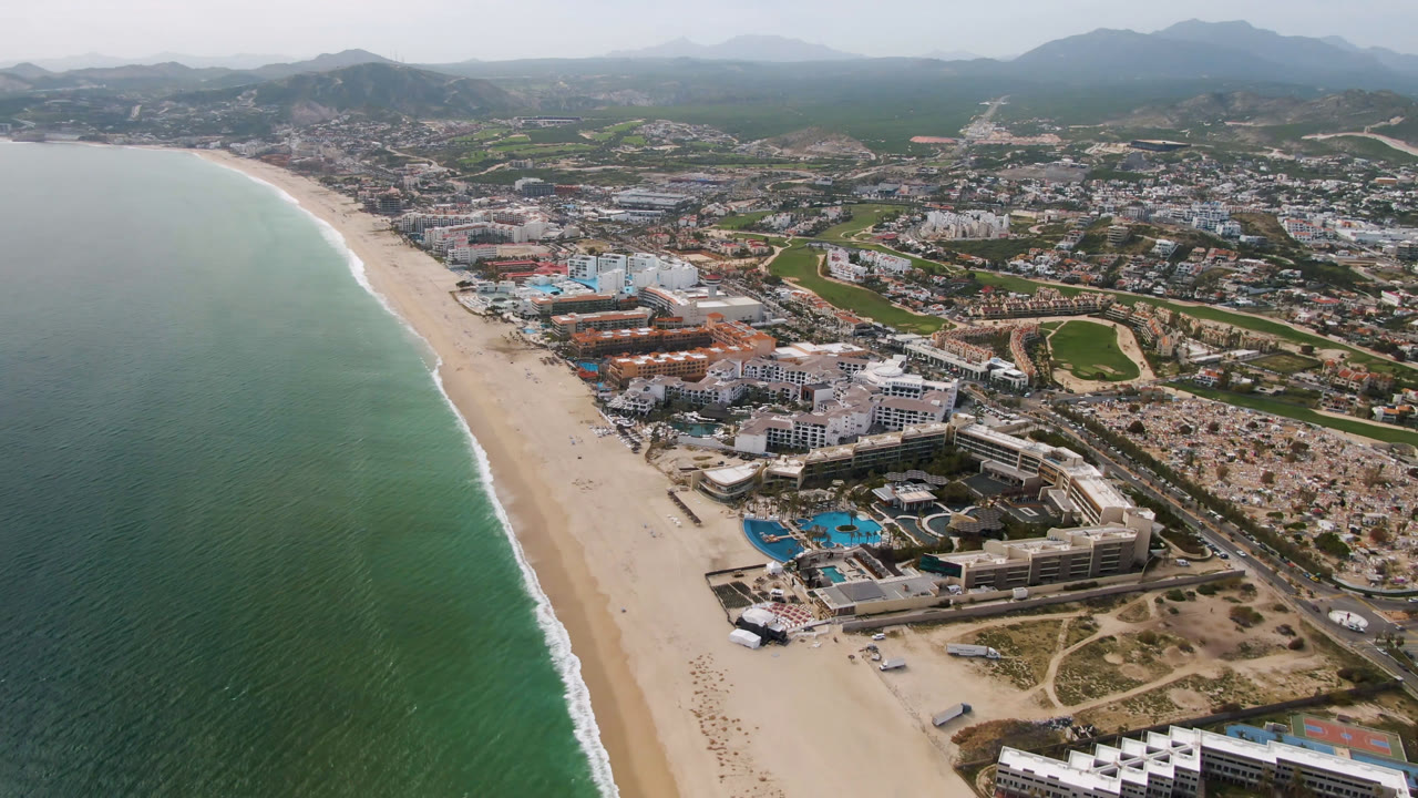 Aerial frame over San Jose Del Cabo showing turquoise ocean at left, a long sandy beach, beachfront resort buildings, inland roads, neighborhoods, golf-course greenery, hazy mountains, and no visible people.