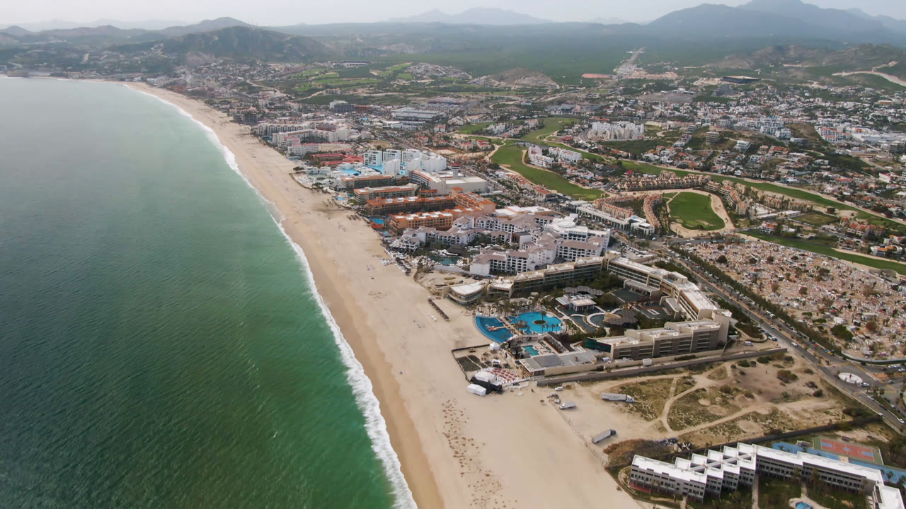 Aerial view in San Jose Del Cabo with the shoreline running diagonally, green-blue water on the left, hotel and resort structures near the beach, inland city blocks, roads, golf-course areas, and no visible people.