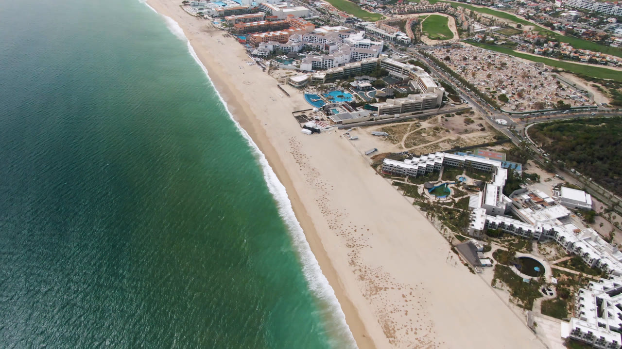 Opening aerial frame over San Jose del Cabo, Baja California Sur, with turquoise ocean on the left, a diagonal sandy beach, resort buildings and pools near the shore, and no visible people.