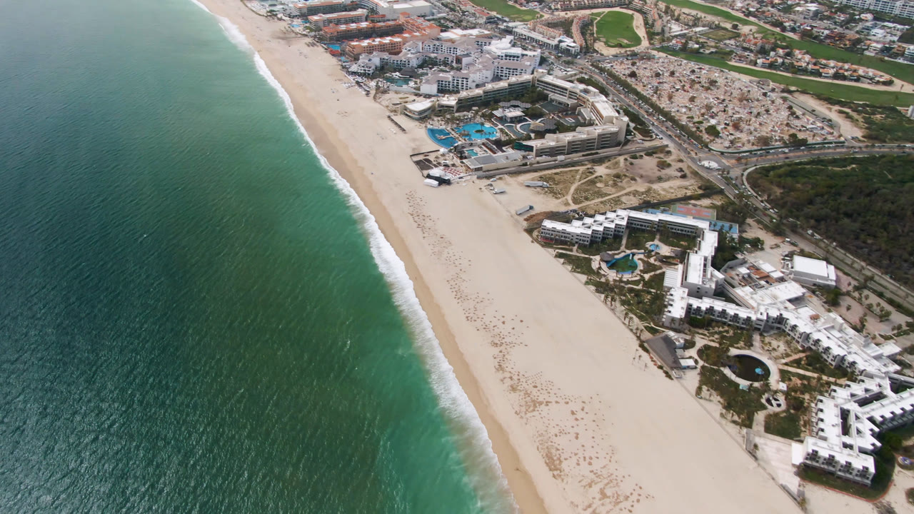 Aerial frame in San Jose del Cabo showing the beach and white surf line beside resort complexes, blue pools, roadways, and inland construction areas, with no visible people.