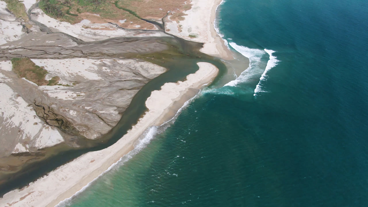Aerial view over San Jose del Cabo showing a pale sandbar between dark inlet water and blue ocean, with white surf on the shoreline and no visible people.