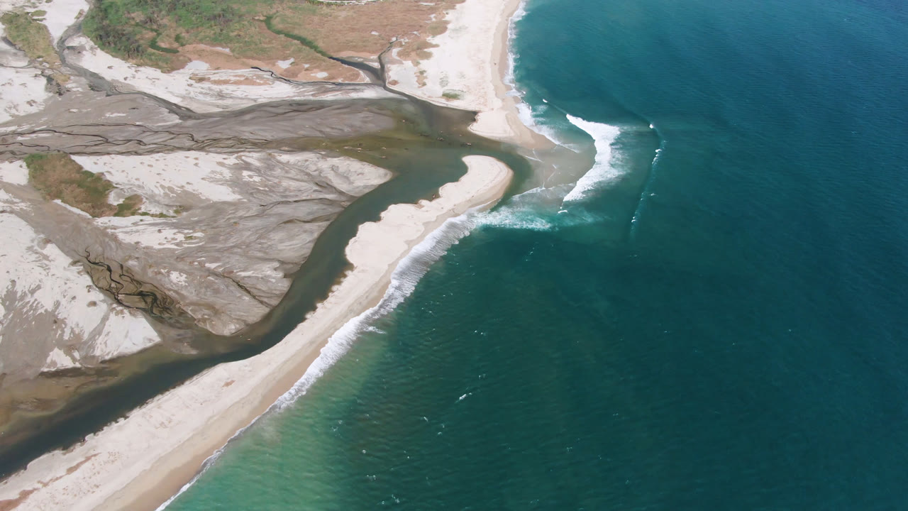 High aerial frame over San Jose del Cabo showing a pale sandbar between a dark inlet and teal ocean, with white surf breaking along the shore and no visible people.