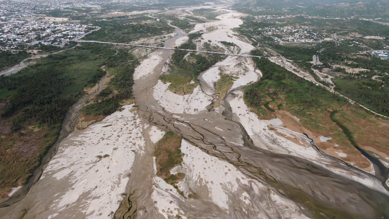 Aerial frame over a pale braided riverbed in San Jose del Cabo, Baja California Sur, with green valley edges, distant roads and buildings, and no visible people.