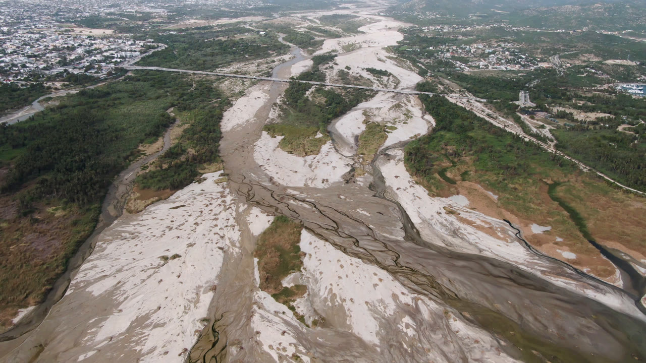 High aerial view in San Jose del Cabo, Baja California Sur, with pale sand channels curving through the valley, darker water traces, wooded slopes, distant roads, and no visible people.