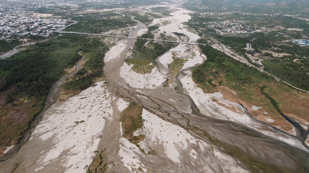 Drone view of a wide dry river wash with roads and buildings beyond.
