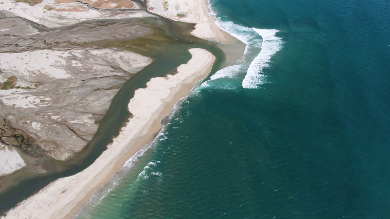 Aerial frame over San Jose del Cabo showing a pale sand spit curving between darker lagoon water and teal ocean, with white surf along the beach and no visible people.