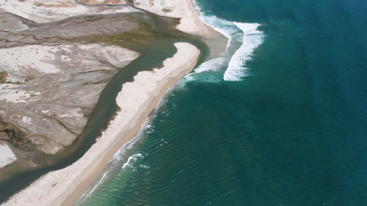 Aerial view in San Jose del Cabo with the long sand bar centered between shallow tidal flats and ocean water, while white waves break near the upper shoreline and no visible people appear.